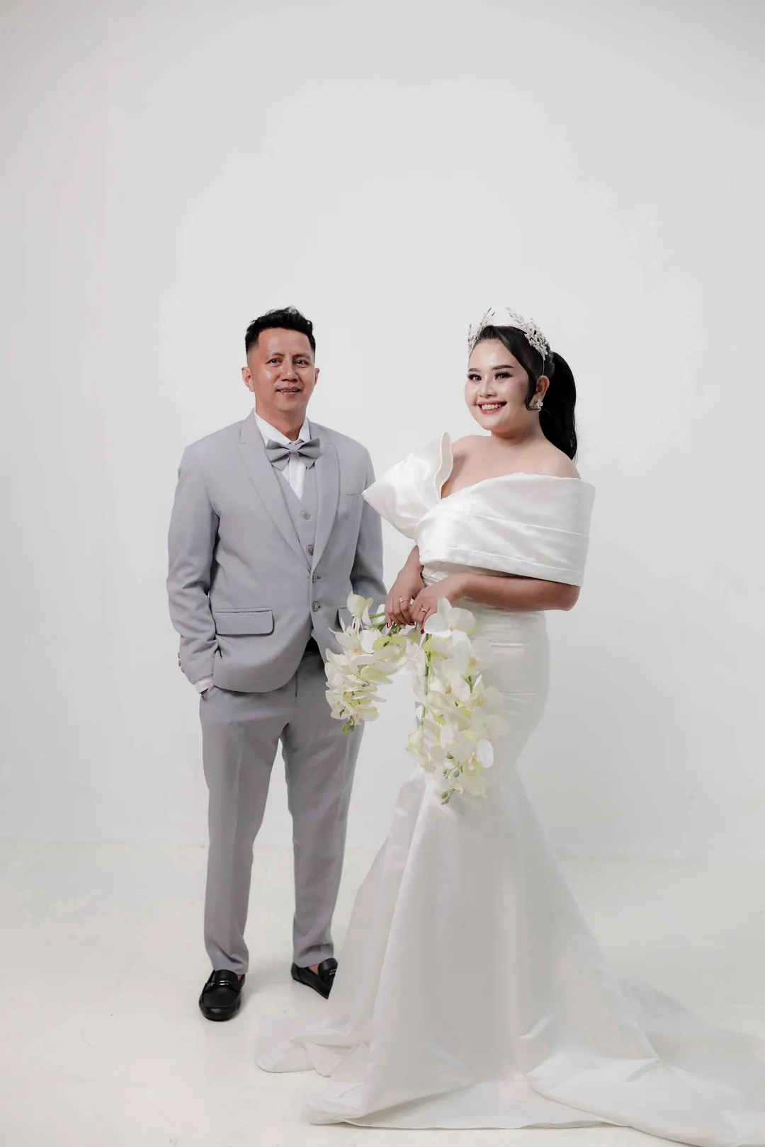 Ryandi berjas abu-abu dan Maria bergaun putih memegang buket anggrek di studio